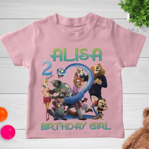 May include: Pink short-sleeved t-shirt featuring a colorful graphic of animated characters, the name "ALISA", the number "2", and the text "BIRTHDAY GIRL". The design includes a blue snake and various animal figures.