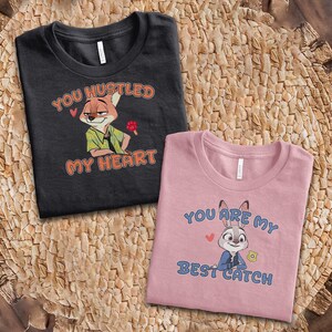 May include: A black t-shirt featuring a cartoon fox and the text "YOU HUSTLED MY HEART" in orange, alongside a pink t-shirt featuring a cartoon rabbit and the text "YOU ARE MY BEST CATCH" in blue, both with small red heart accents.