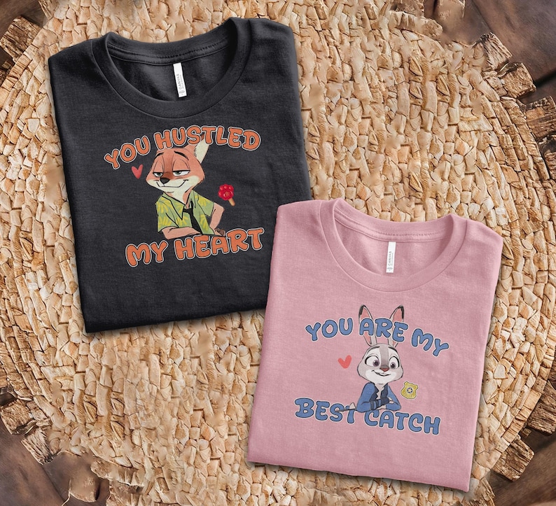 May include: A black t-shirt featuring a cartoon fox and the text "YOU HUSTLED MY HEART" in orange, alongside a pink t-shirt featuring a cartoon rabbit and the text "YOU ARE MY BEST CATCH" in blue, both with small red heart accents.