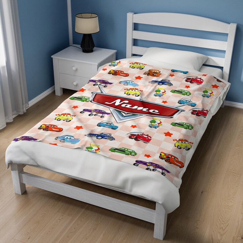 May include: A children's blanket featuring a repeating pattern of cartoon cars in various colors on a pink and white checkered background. A red sign with the name "Name" is centered on the blanket. The blanket is on a white bed.