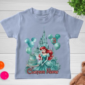 以下が含まれることがあります： 水色のTシャツ。お城、風船、ケーキなど、プリンセスをテーマにしたデザイン。数字の3は赤色で、「Custom Name」の文字は筆記体です。プリンセスは赤い髪で、ターコイズブルーのドレスを着ています。