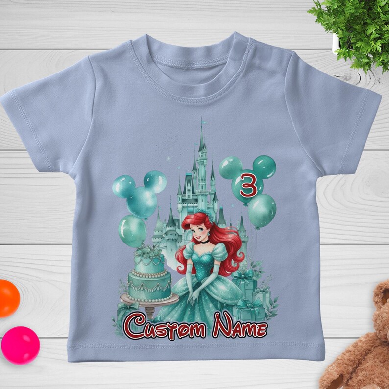 以下が含まれることがあります： 水色のTシャツ。お城、風船、ケーキなど、プリンセスをテーマにしたデザイン。数字の3は赤色で、「Custom Name」の文字は筆記体です。プリンセスは赤い髪で、ターコイズブルーのドレスを着ています。