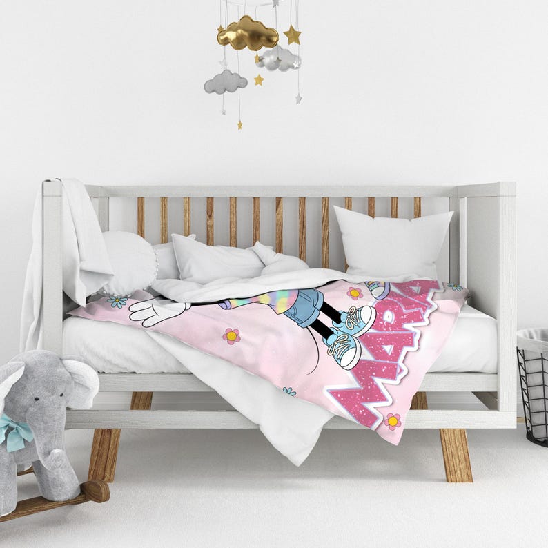 Op de afbeelding: Een roze babydeken met een Minnie Mouse-ontwerp. De deken heeft een grote afbeelding van Minnie Mouse en het woord "Minnie" in roze letters. De deken ligt op een wit babybedje met een grijs olifantenspeelgoed.