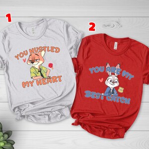 May include: A light gray t-shirt featuring a cartoon fox and the text "YOU HUSTLED MY HEART" in orange, alongside a red t-shirt featuring a cartoon rabbit and the text "YOU ARE MY BEST CATCH" in blue, both with short sleeves and crew necklines.