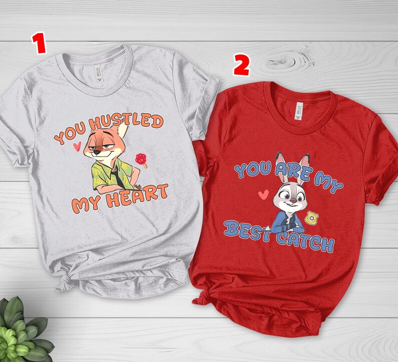 May include: A light gray t-shirt featuring a cartoon fox and the text "YOU HUSTLED MY HEART" in orange, alongside a red t-shirt featuring a cartoon rabbit and the text "YOU ARE MY BEST CATCH" in blue, both with short sleeves and crew necklines.