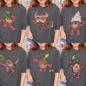 Puede incluir: Seis camisetas de color gris antracita con diferentes dise&ntilde;os de monstruos de dibujos animados. Cada monstruo tiene un cuerpo marr&oacute;n, extremidades rosas y varios accesorios como lanzas, garrotes y tocados. Los dise&ntilde;os est&aacute;n centrados en las camisetas.