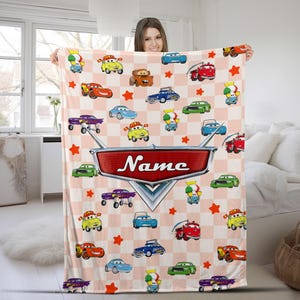 May include: A pink and white checkered blanket featuring cartoon cars and stars. The blanket has a large red emblem with the word "Name" in white. The blanket is held up by a person.