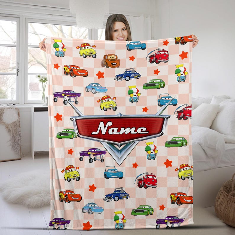 May include: A pink and white checkered blanket featuring cartoon cars and stars. The blanket has a large red emblem with the word "Name" in white. The blanket is held up by a person.