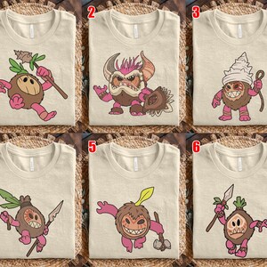 Puede incluir: Camisetas beige con varios dise&ntilde;os de personajes de dibujos animados. Cada camiseta presenta un personaje diferente con un cuerpo marr&oacute;n, extremidades rosas y un accesorio &uacute;nico, como una lanza o un escudo. Los personajes tienen caras expresivas.