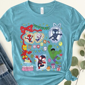 May include: A light blue t-shirt featuring a colorful graphic print of various superheroes, including Iron Man, Hulk, and Spider-Man characters. The design includes a birthday cake, gift boxes, balloons, bunting, and the text NAME 5.