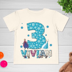 May include: Cream-colored t-shirt featuring a large blue number 3 filled with snowflakes. The name "VIVIAN" is printed below the number. Cartoon images of characters are also on the shirt. A birthday-themed shirt.