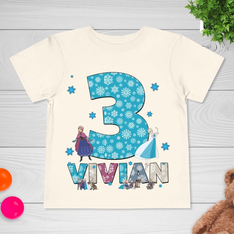 May include: Cream-colored t-shirt featuring a large blue number 3 filled with snowflakes. The name "VIVIAN" is printed below the number. Cartoon images of characters are also on the shirt. A birthday-themed shirt.