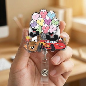 Op de afbeelding: Een kleurrijke badge reel met tekenfilmauto's Mater en Lightning McQueen, met Mickey Mouse oren, en een cluster van Mickey Mouse-vormige ballonnen. De badge reel heeft een doorzichtig plastic intrekbaar koord en een knopclip.