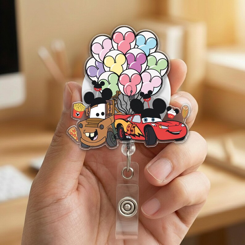 Op de afbeelding: Een kleurrijke badge reel met tekenfilmauto's Mater en Lightning McQueen, met Mickey Mouse oren, en een cluster van Mickey Mouse-vormige ballonnen. De badge reel heeft een doorzichtig plastic intrekbaar koord en een knopclip.
