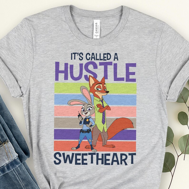 May include: A light gray crew neck t-shirt featuring a graphic of Judy Hopps and Nick Wilde. The text reads IT'S CALLED A HUSTLE SWEETHEART. The characters are positioned over horizontal stripes of purple, green, blue, pink, and orange colors.