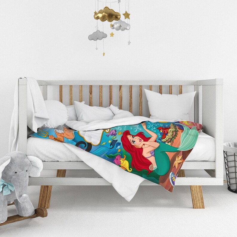 May include: A white crib blanket featuring a colorful underwater scene with a red-haired mermaid, a king with a trident, and various sea creatures including fish and a crab, all set against a vibrant blue ocean background with bubbles.