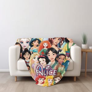 Op de afbeelding: Een zachte, kleurrijke deken met een collage van geanimeerde prinsessen. De deken is over een witte bank gedrapeerd en bevat de naam "Alice" in een gestileerd lettertype. Het ontwerp toont verschillende prinsessen in levendige kleuren.
