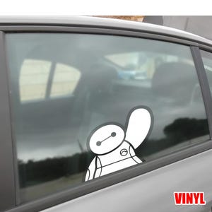 May include: A white vinyl decal of Baymax from Big Hero 6, affixed to a car window. The decal features Baymax's head and upper body, with a simple, clean design. The word "VINYL" is printed in red in the lower right corner.