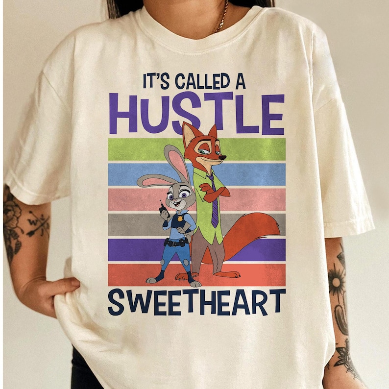 May include: A cream-colored short-sleeve t-shirt featuring a graphic of a rabbit and a fox. The text "IT'S CALLED A HUSTLE SWEETHEART" is printed in dark blue. The background of the graphic consists of horizontal stripes in green, blue, pink, gray, purple, and orange.