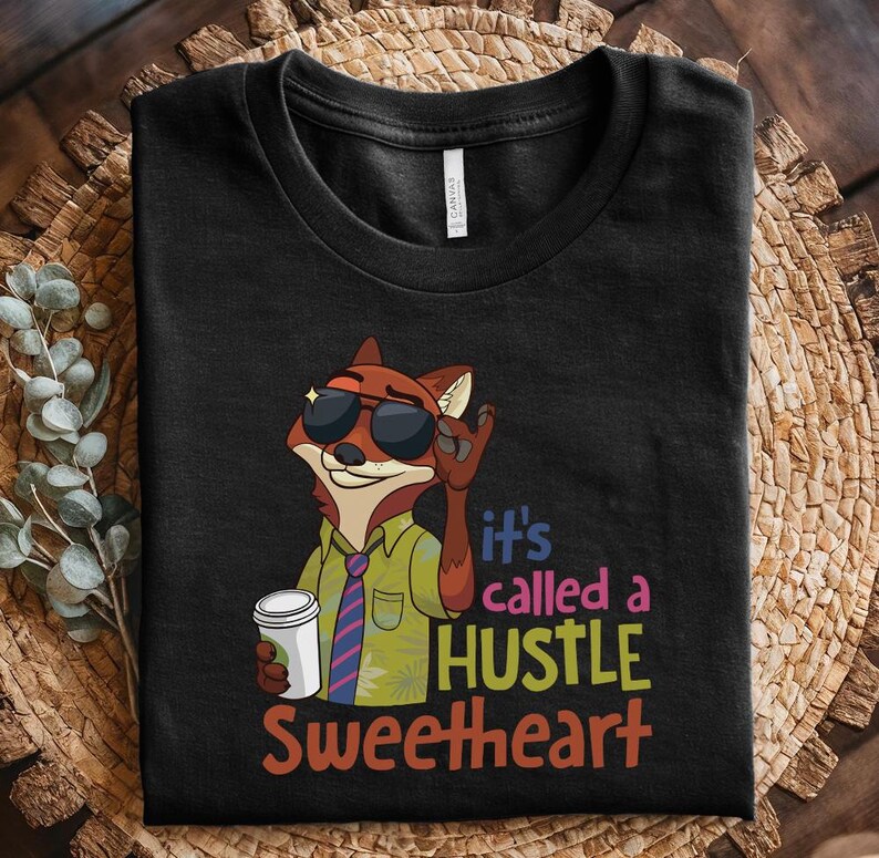 Op de afbeelding: Zwart t-shirt met een cartoonvos met een zonnebril en een koffiekopje. De vos maakt een handgebaar. De tekst op het shirt luidt "It's called a HUSTLE Sweetheart" in kleurrijke letters.