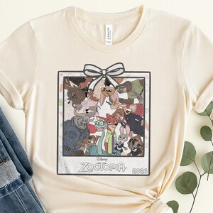 Puede incluir: Camiseta color crema con un gr&aacute;fico enmarcado de personajes de la pel&iacute;cula de Disney "Zootopia". El gr&aacute;fico incluye un lazo en la parte superior y el t&iacute;tulo de la pel&iacute;cula y el a&ntilde;o, "2026", en la parte inferior. La camiseta es de un material suave.