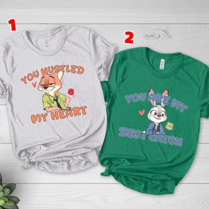 May include: A light gray t-shirt featuring a cartoon fox and the text "YOU HUSTLED MY HEART" in orange, alongside a green t-shirt featuring a cartoon rabbit and the text "YOU ARE MY BEST CATCH" in blue, both with small red heart graphics.