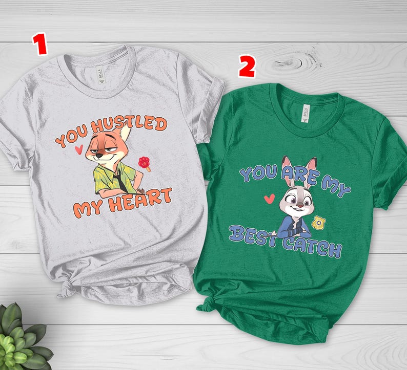 May include: A light gray t-shirt featuring a cartoon fox and the text "YOU HUSTLED MY HEART" in orange, alongside a green t-shirt featuring a cartoon rabbit and the text "YOU ARE MY BEST CATCH" in blue, both with small red heart graphics.