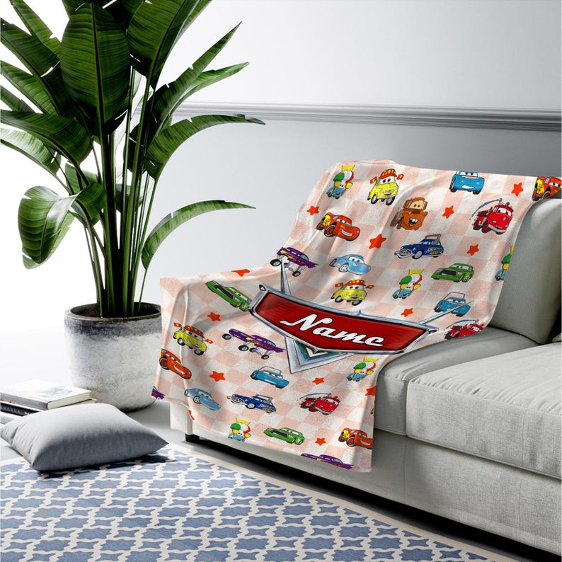May include: A soft, light pink blanket with a repeating pattern of cartoon cars and red stars. The blanket features a red banner with the word "Name" on it. The blanket is draped over a white couch.
