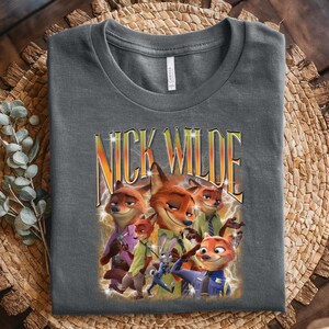 May include: Dark gray t-shirt featuring a graphic of Nick Wilde from Zootopia. The design includes multiple images of the fox character with the text "NICK WILDE" in a yellow and orange gradient font with lightning bolt accents.