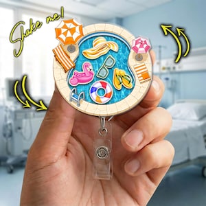May include: A round badge reel featuring a pool scene with a flamingo float, sunglasses, and a beach hat. The badge reel has a clear plastic clip and the words "Shake me!" in a yellow script.