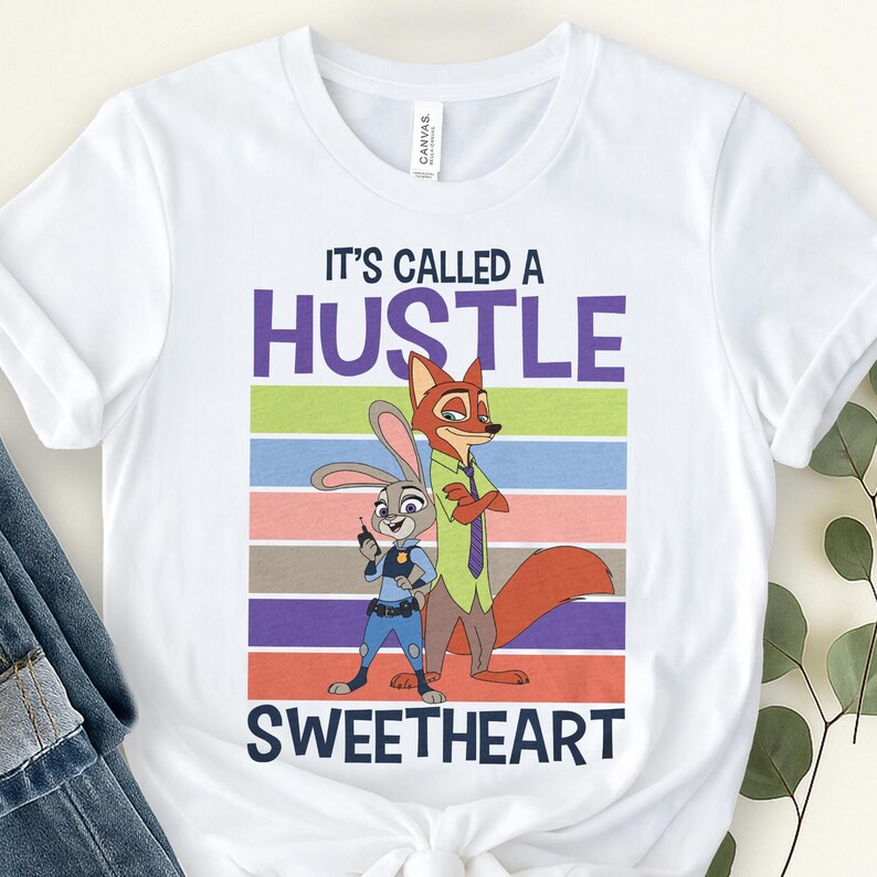 May include: A white short-sleeved t-shirt featuring a graphic of Judy Hopps and Nick Wilde from Zootopia. The text reads IT'S CALLED A HUSTLE SWEETHEART. The design includes horizontal stripes in green, blue, pink, brown, and purple behind the characters.