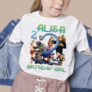 May include: White t-shirt featuring a colorful graphic of characters from the movie Zootopia, with the name "ALISA", the number "2", and the text "BIRTHDAY GIRL" in green and blue. The shirt is a children's apparel item.