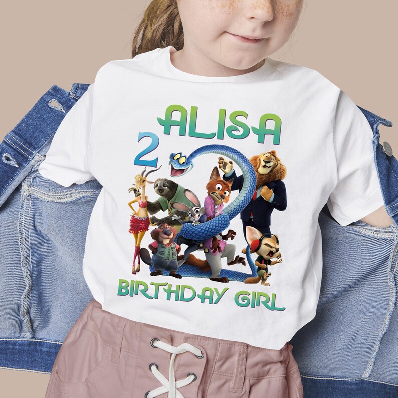 May include: White t-shirt featuring a colorful graphic of characters from the movie Zootopia, with the name "ALISA", the number "2", and the text "BIRTHDAY GIRL" in green and blue. The shirt is a children's apparel item.