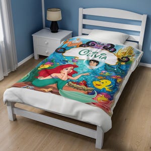 May include: A white blanket featuring a colorful underwater scene with the character Ariel, Flounder, and other sea creatures. The design includes a central white cloud shape with the name Olivia printed in green script text on the fabric.