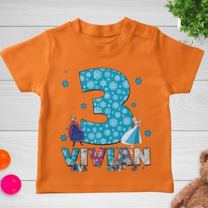 May include: Orange short-sleeved t-shirt featuring a large blue number 3 filled with white snowflakes. The name "VIVIAN" is printed below the number, with images of cartoon characters. Small blue snowflakes are scattered around the number.