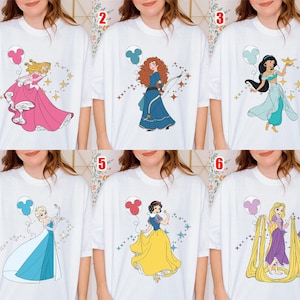 May include: White t-shirts featuring cartoon illustrations of Disney princesses. Each shirt showcases a different princess, including Aurora in a pink gown, Merida in a blue dress, and Jasmine in a turquoise outfit. Each princess is accompanied by a Mickey Mouse balloon.