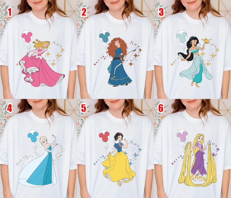 May include: White t-shirts featuring cartoon illustrations of Disney princesses. Each shirt showcases a different princess, including Aurora in a pink gown, Merida in a blue dress, and Jasmine in a turquoise outfit. Each princess is accompanied by a Mickey Mouse balloon.
