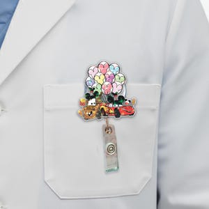 Op de afbeelding: Een intrekbare badge reel met de stripfiguren Mater en Lightning McQueen uit de film Cars, die kleurrijke ballonnen vasthouden. De reel is bevestigd aan een doorzichtige plastic clip met een knop.