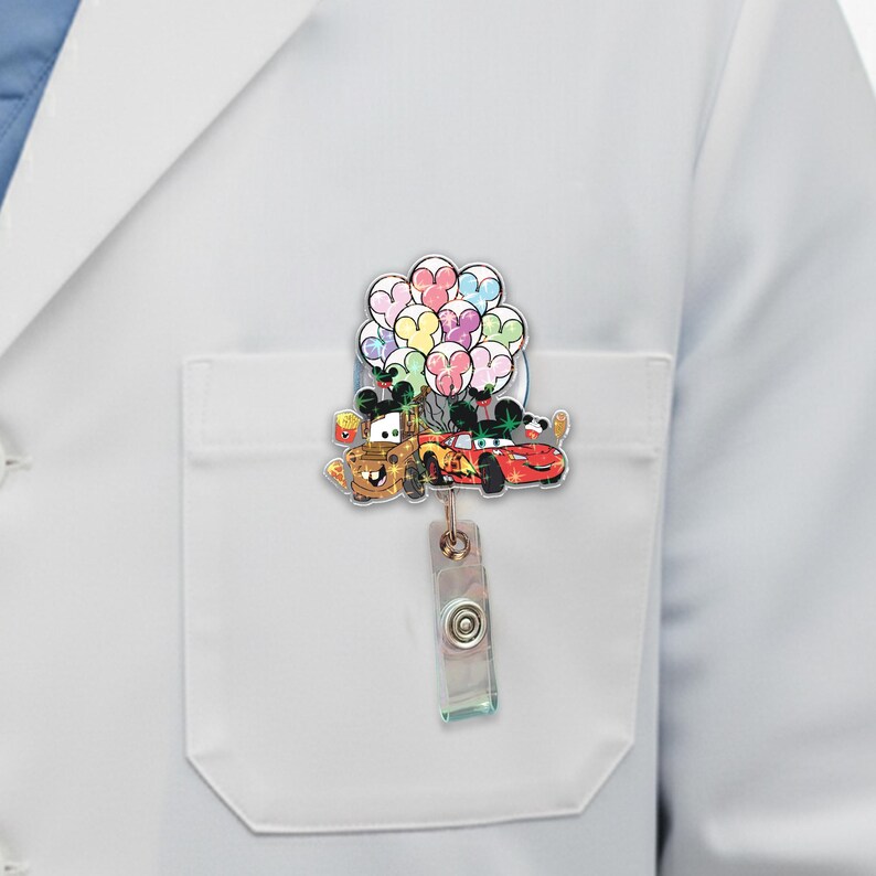 Op de afbeelding: Een intrekbare badge reel met de stripfiguren Mater en Lightning McQueen uit de film Cars, die kleurrijke ballonnen vasthouden. De reel is bevestigd aan een doorzichtige plastic clip met een knop.