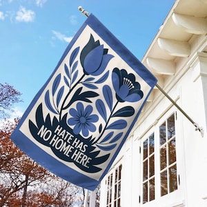 May include: A rectangular garden flag with a blue border and a cream-colored center. The flag features a floral design in shades of blue, with the text "HATE HAS NO HOME HERE."