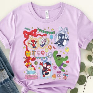 May include: A light purple short-sleeve t-shirt featuring a colorful graphic print of various superheroes, a birthday cake, presents, balloons, and stars. The design includes the text "NAME" and the number "5" surrounded by festive party elements.