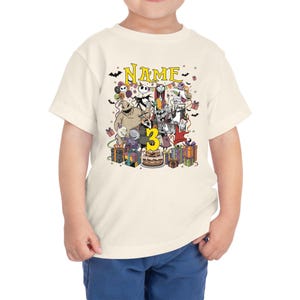 May include: Cream-colored toddler t-shirt featuring a colorful graphic of characters from The Nightmare Before Christmas. The graphic includes the word "Name" above the characters and the number "3". Presents and a birthday cake are also included.