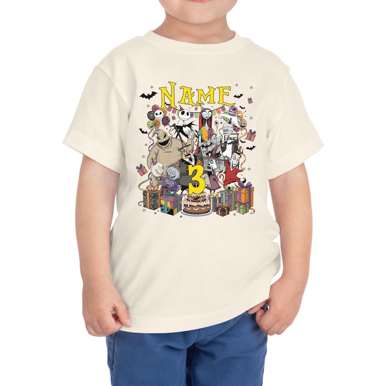 May include: Cream-colored toddler t-shirt featuring a colorful graphic of characters from The Nightmare Before Christmas. The graphic includes the word "Name" above the characters and the number "3". Presents and a birthday cake are also included.