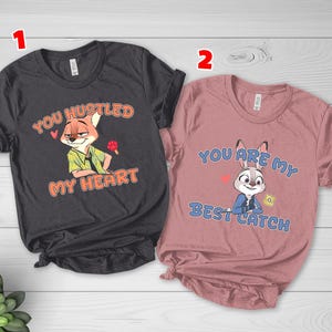 May include: A dark gray t-shirt featuring a cartoon fox and the text YOU HUSTLED MY HEART, and a dusty rose t-shirt featuring a cartoon rabbit and the text YOU ARE MY BEST CATCH. Both shirts have short sleeves and crew necklines with printed graphics.