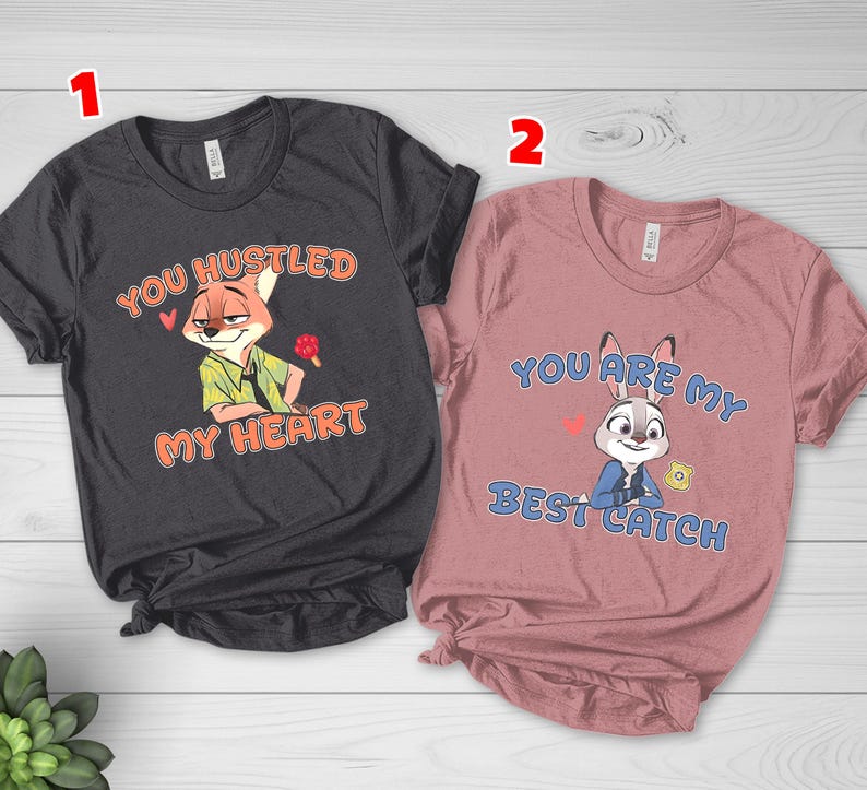 May include: A dark gray t-shirt featuring a cartoon fox and the text YOU HUSTLED MY HEART, and a dusty rose t-shirt featuring a cartoon rabbit and the text YOU ARE MY BEST CATCH. Both shirts have short sleeves and crew necklines with printed graphics.