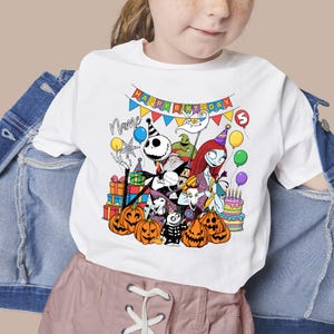 May include: White t-shirt featuring a colorful graphic of characters from The Nightmare Before Christmas, including Jack Skellington, Sally, and Oogie Boogie. The design includes pumpkins, balloons, and a birthday banner that says "Happy Birthday".