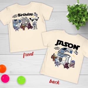 May include: Two cream-colored t-shirts with a Star Wars theme. One shirt says "Birthday Boy" with character graphics. The other shirt says "Jason" with character graphics. The shirts are on a light wood surface.