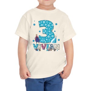 May include: A cream-colored toddler t-shirt featuring a large turquoise number "3" filled with snowflakes. The name "Vivian" is below the number. The shirt has images of two animated characters from the movie Frozen.