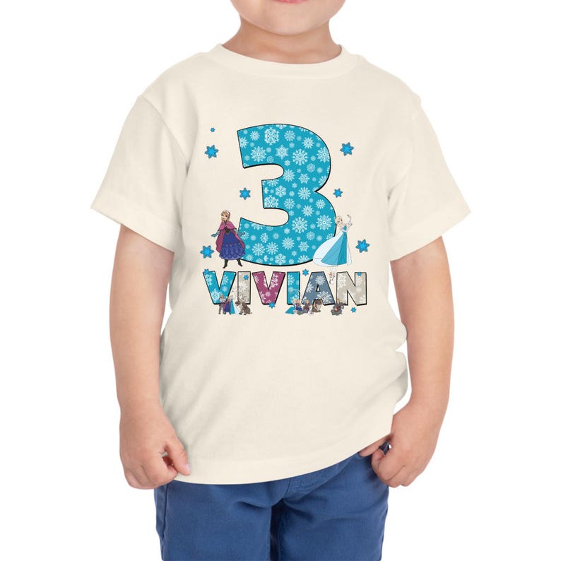May include: A cream-colored toddler t-shirt featuring a large turquoise number "3" filled with snowflakes. The name "Vivian" is below the number. The shirt has images of two animated characters from the movie Frozen.