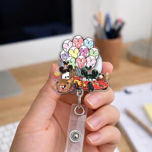 Op de afbeelding: Een doorzichtige badge reel met de tekenfilmfiguren Mater en Lightning McQueen uit de film Cars, met een tros kleurrijke ballonnen. De reel heeft een doorzichtige plastic band en een metalen clip.